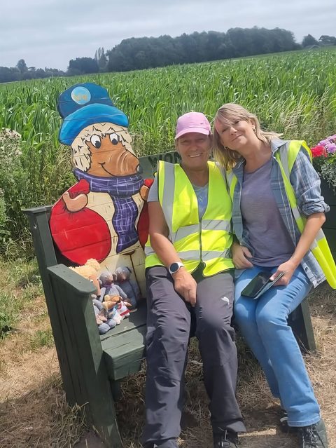 Two volunteers in hi-vis vests sitting together on a green bench beside a painted wooden Womble figure, with a cornfield behind them