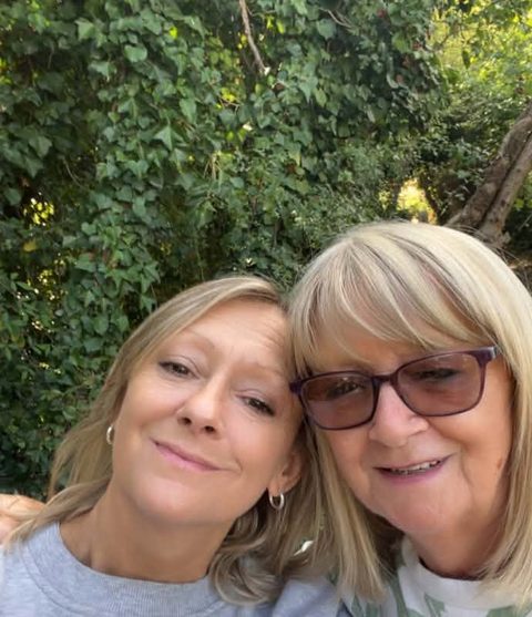 Two smiling women posing close together for a photo, with green leaves behind them